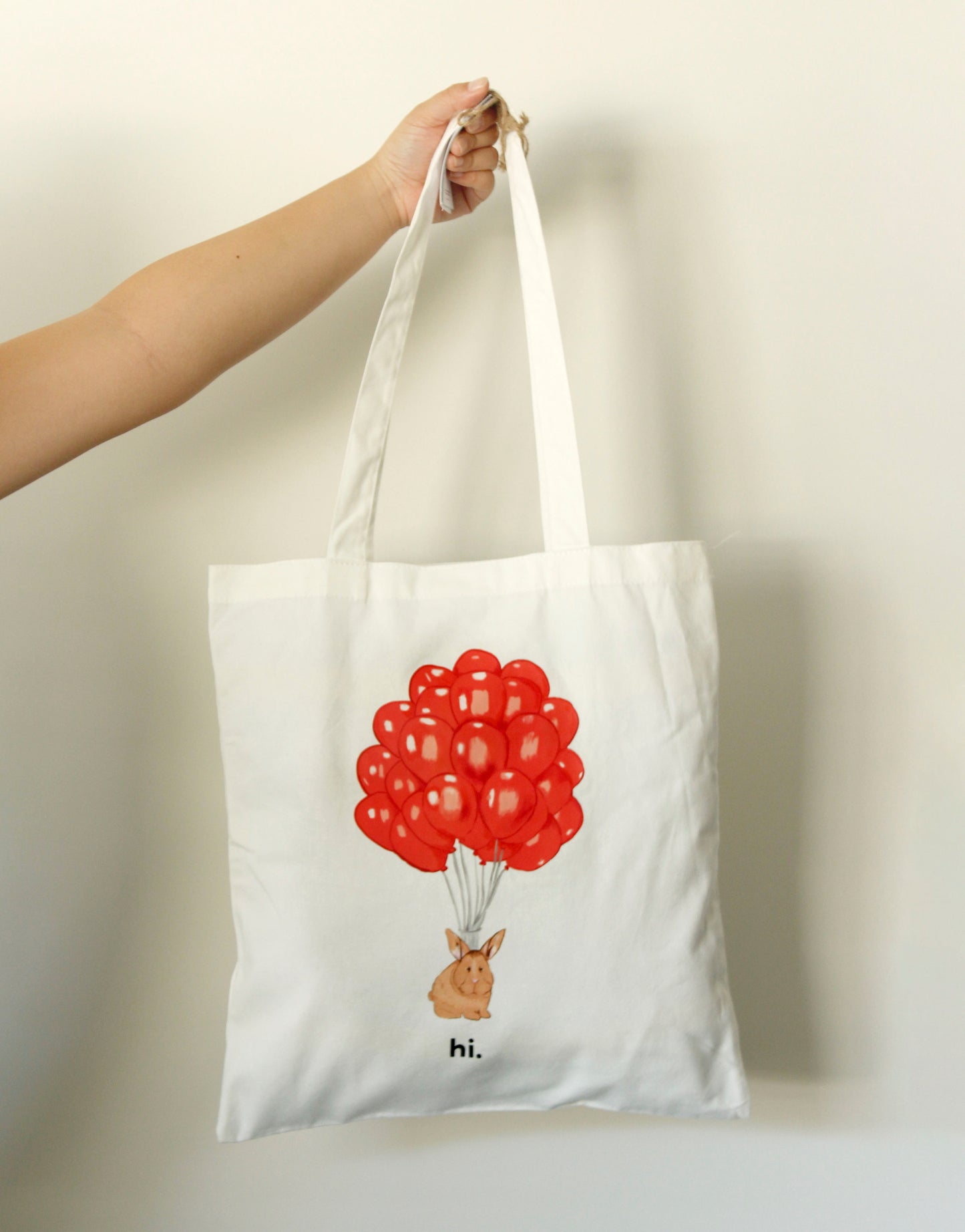 Tote bag with a rabbit holding red balloons and 'hi.' text, held by a hand against a plain background.