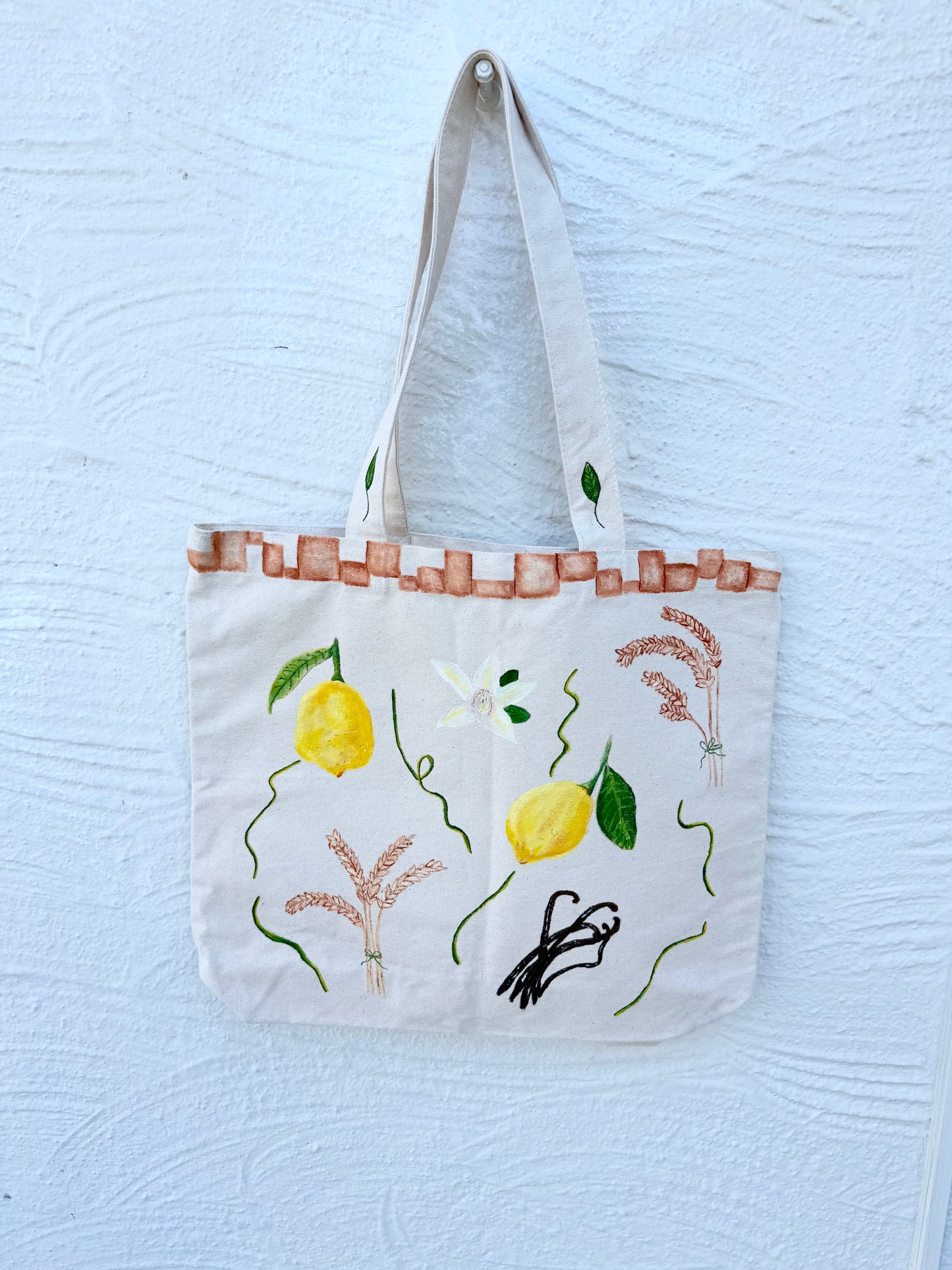 Photo of a hand-painted thick canvased tote bag for sale with vibrant yellow lemons, hand-painted wheat branches, vanilla and terracotta tiles painted on the border of the bag near the handles.