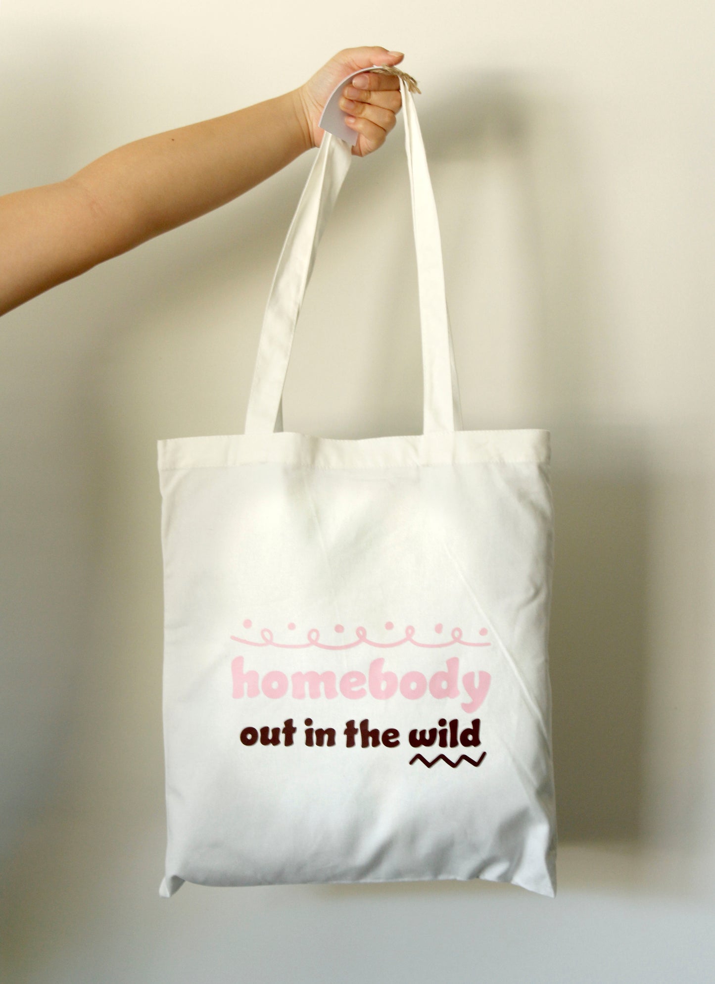 Tote bag with 'homebody out in the wild' text held by a hand against a neutral background