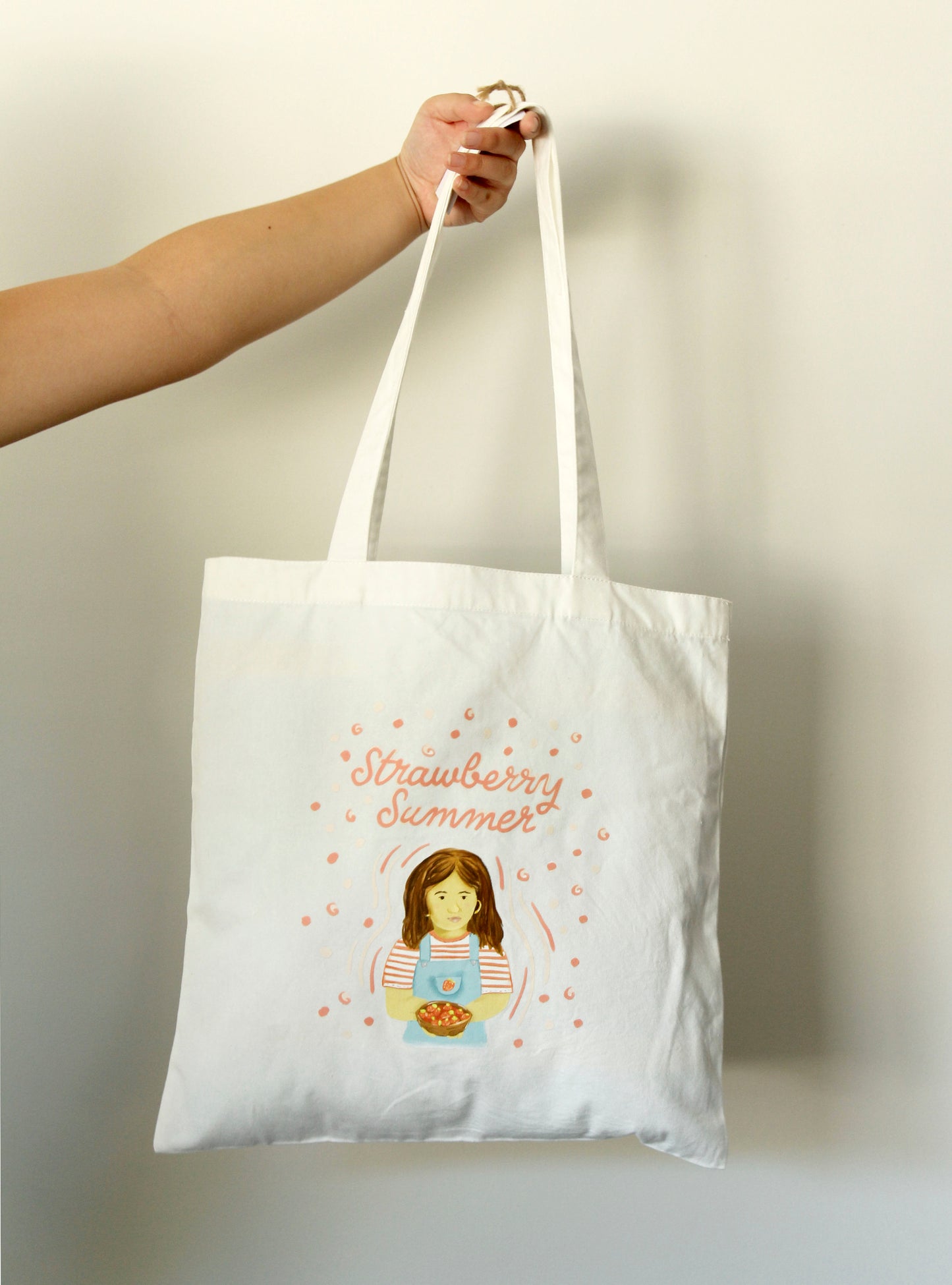 White tote bag with 'Strawberry Summer' design held by a hand against a plain background