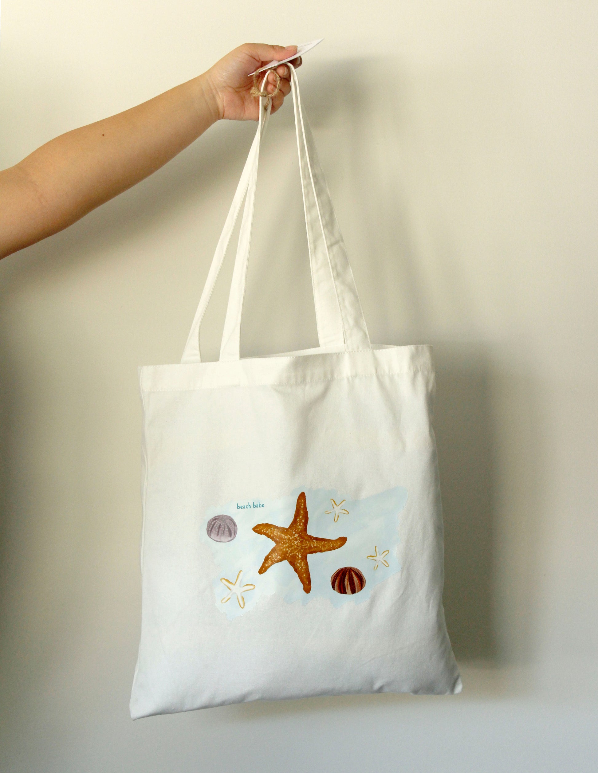 White tote bag with beach-themed designs held by a hand against a plain background