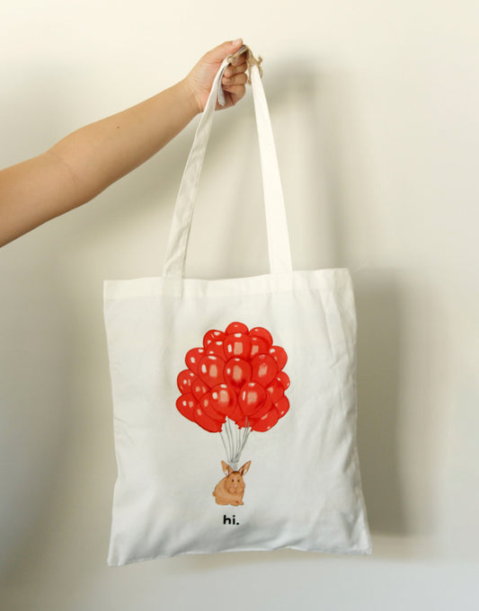 Tote bag with a rabbit holding red balloons and 'hi.' text, held by a hand against a plain background.
