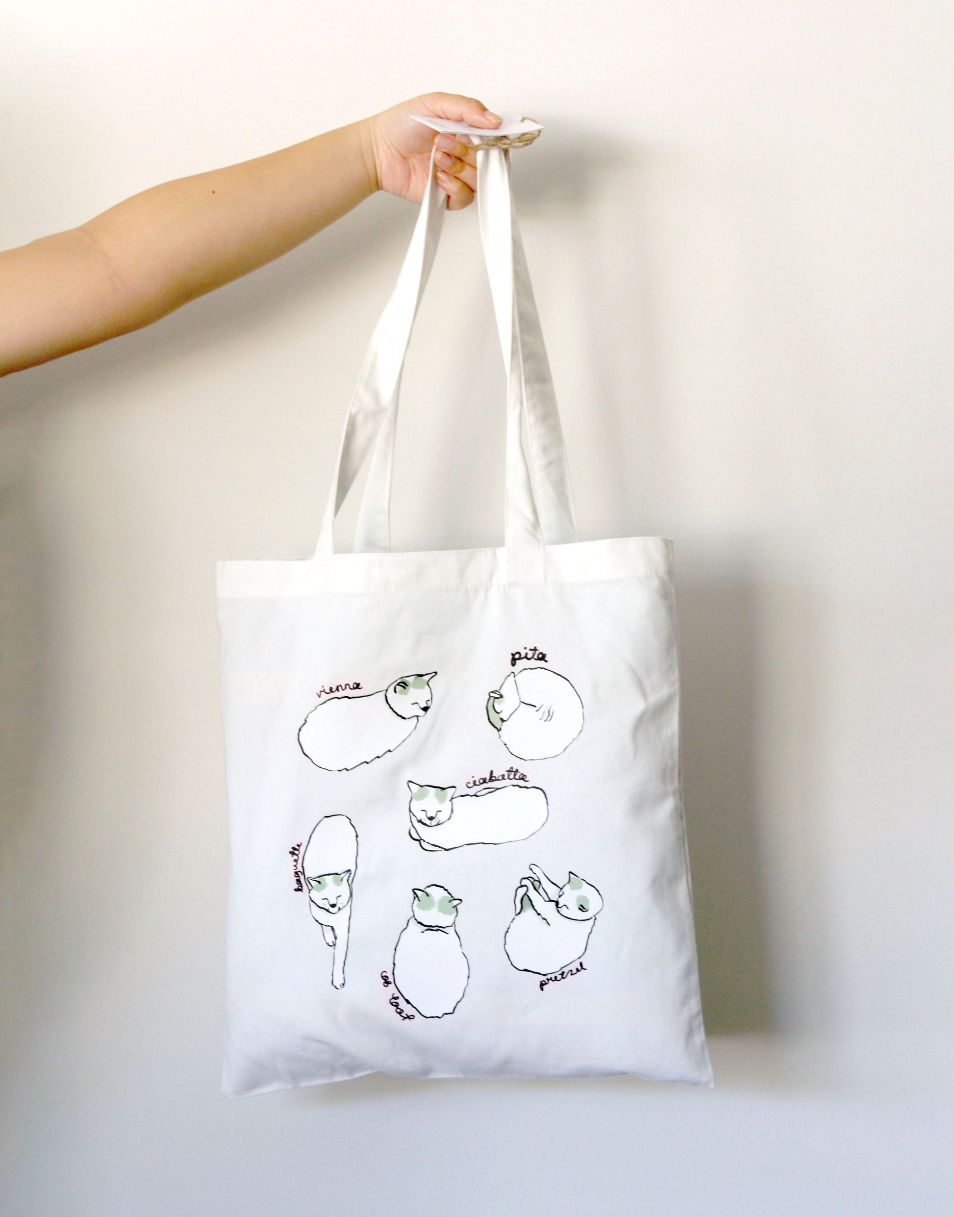 White tote bag with cat loaf illustrations held by a hand against a white background