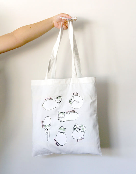 White tote bag with cat loaf illustrations held by a hand against a white background