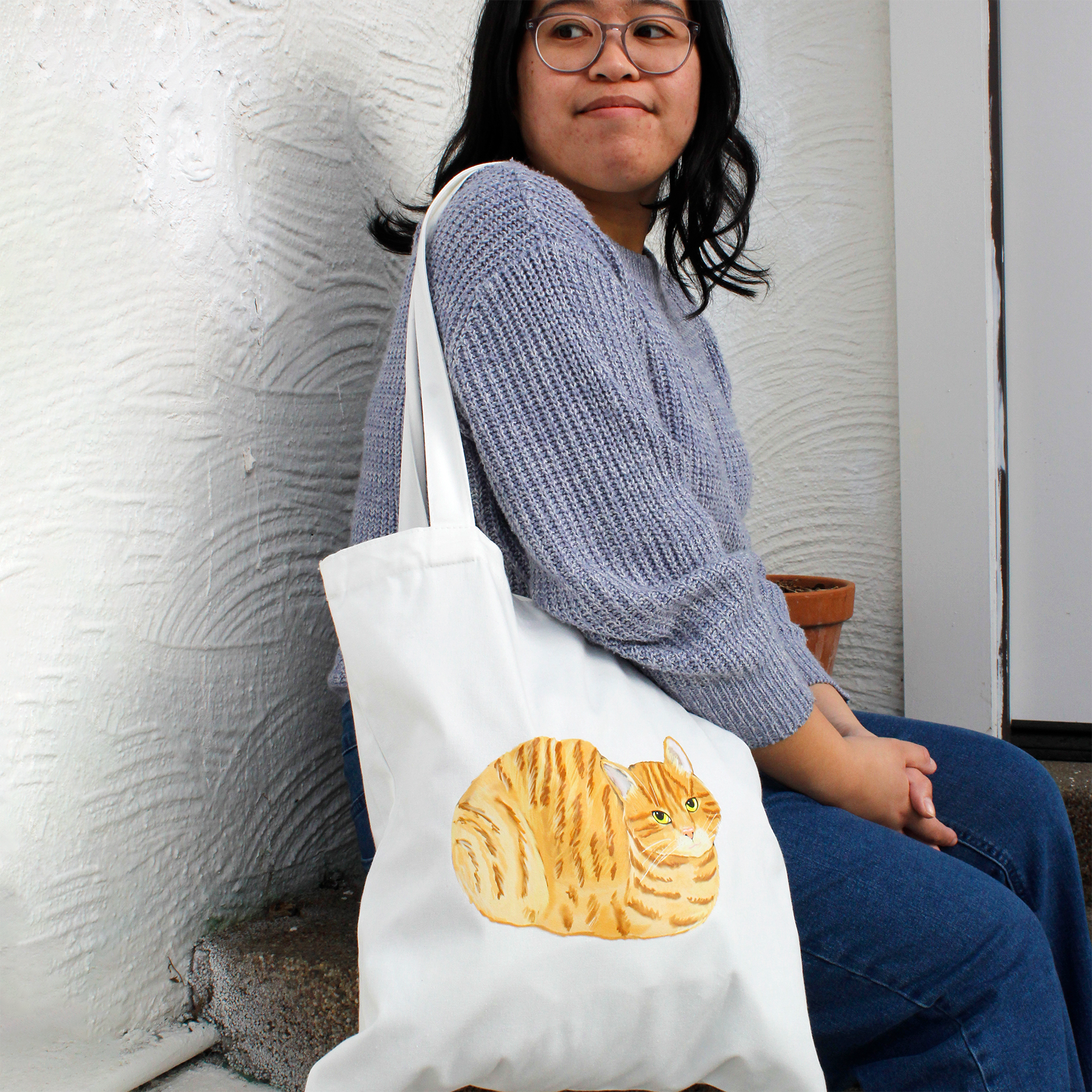 Person holding a white tote bag with a design of a ginger cat loafing on it against a textured wall.