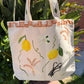 Photo of a hand-painted thick canvased tote bag for sale with vibrant yellow lemons, hand-painted wheat branches, vanilla and terracotta tiles painted on the border of the bag near the handles.