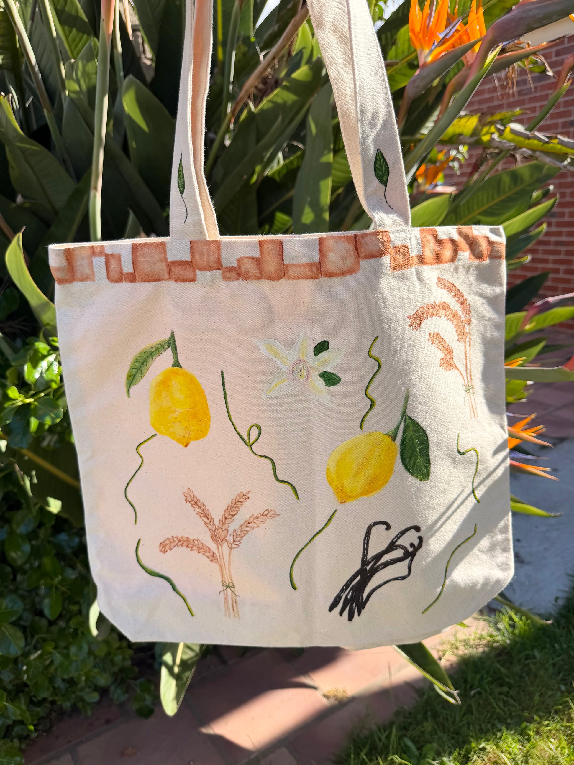 Photo of a hand-painted thick canvased tote bag for sale with vibrant yellow lemons, hand-painted wheat branches, vanilla and terracotta tiles painted on the border of the bag near the handles.