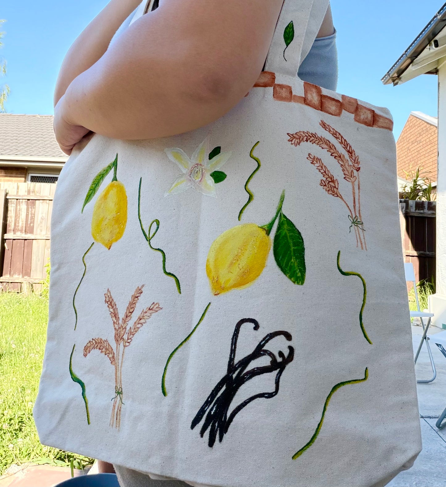 Photo of a hand-painted thick canvased tote bag for sale with vibrant yellow lemons, hand-painted wheat branches, vanilla and terracotta tiles painted on the border of the bag near the handles.