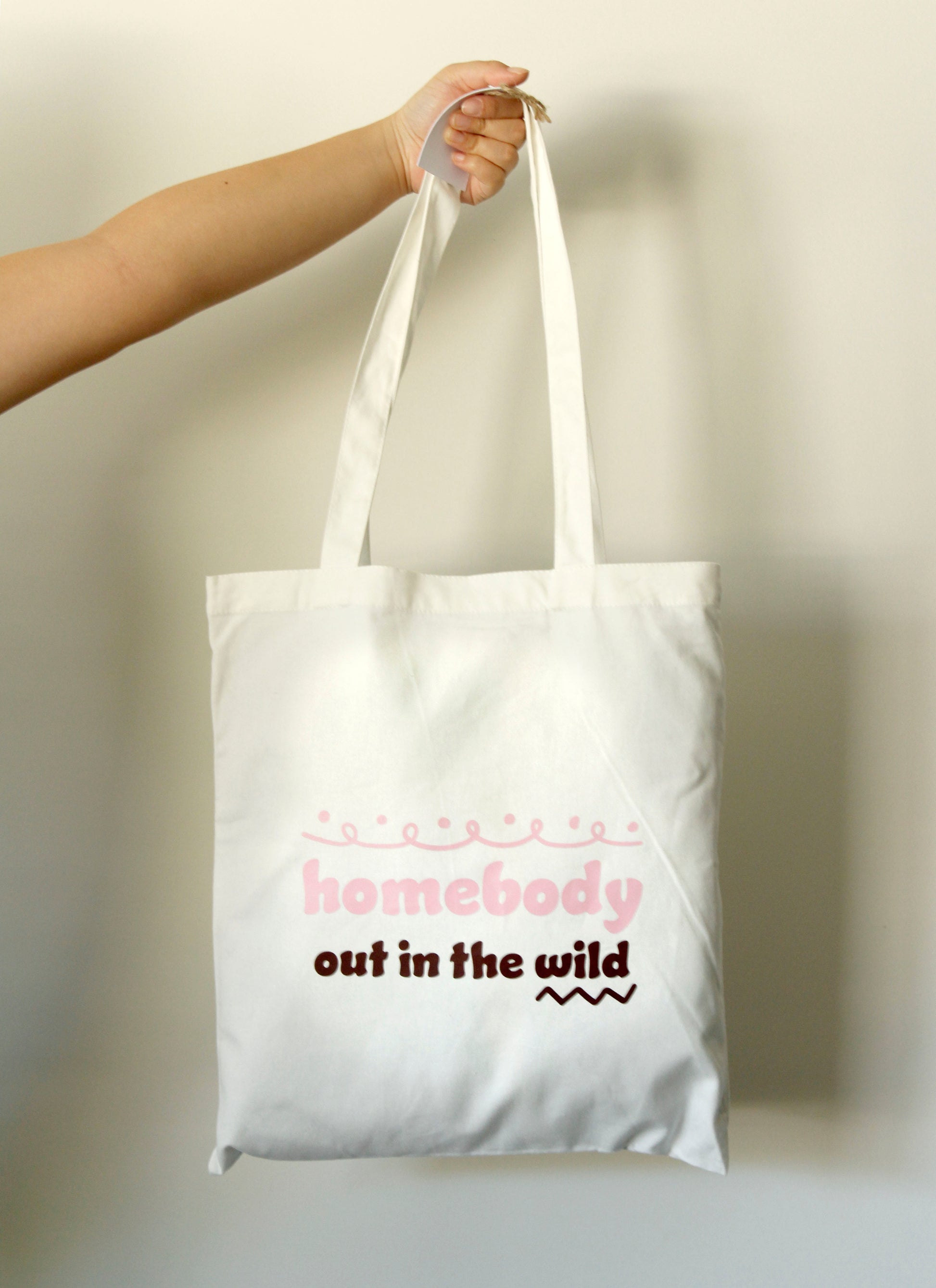 Tote bag with 'homebody out in the wild' text held by a hand against a neutral background