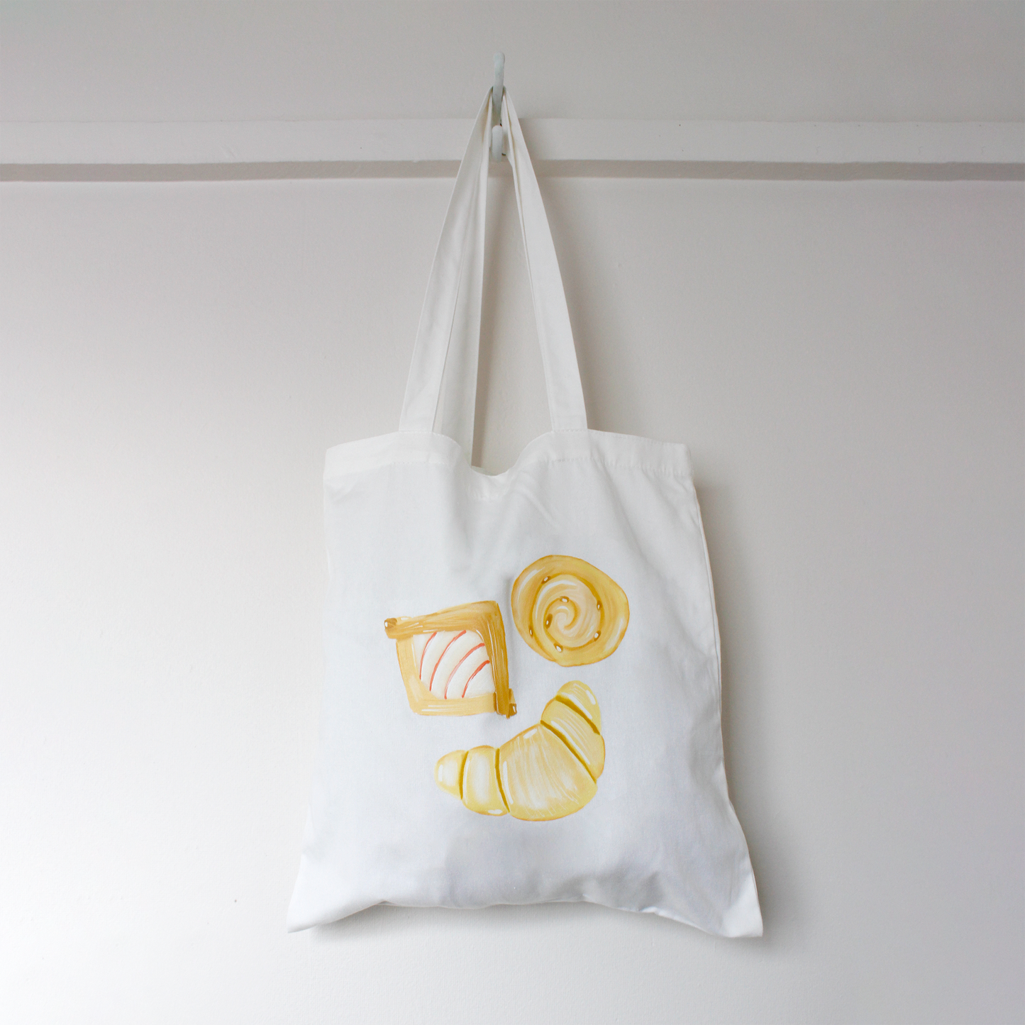 White tote bag with yellow croissant and danish pastries design hanging on a white wall.
