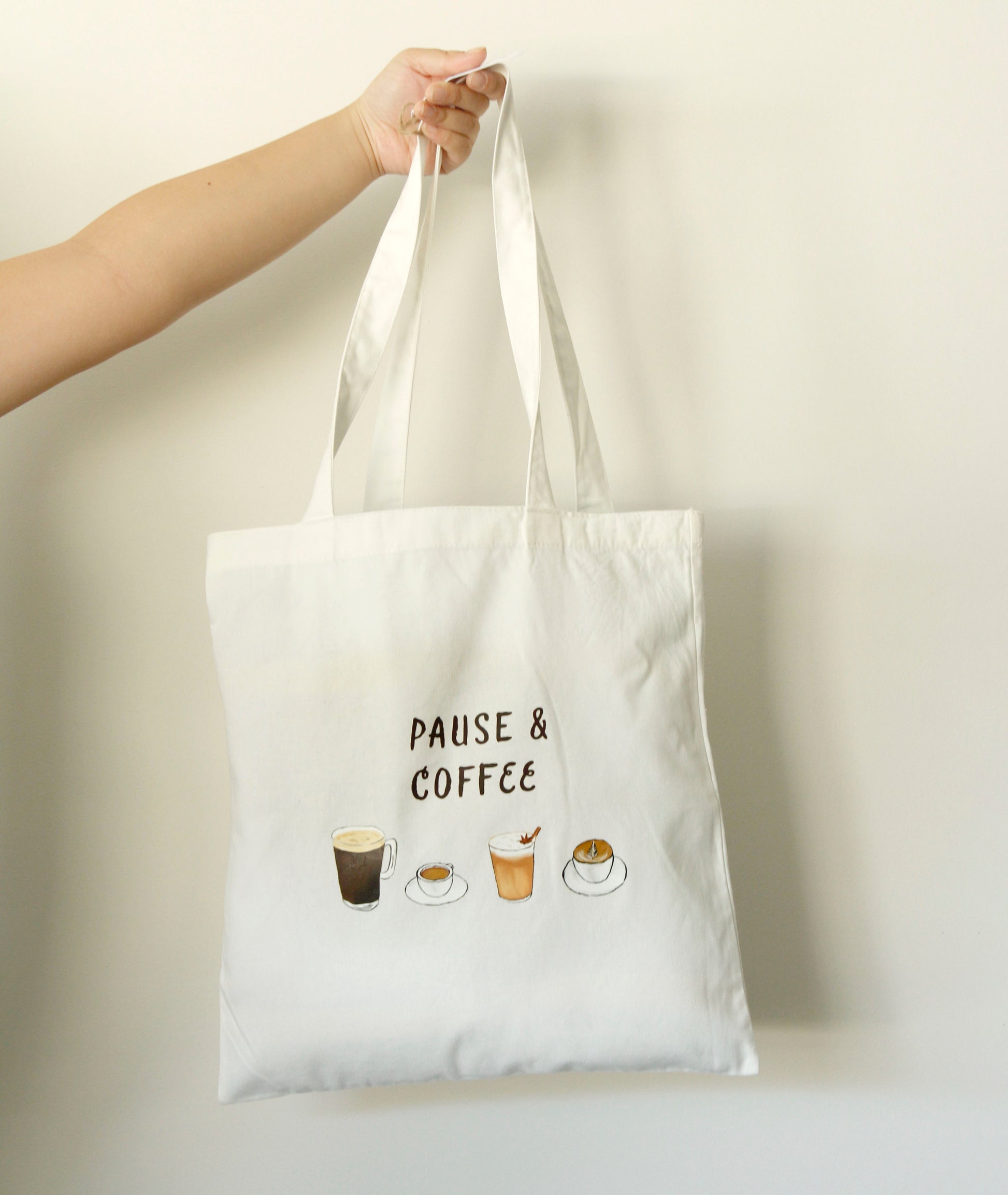 White tote bag with 'Pause & Coffee' text and coffee cup illustrations held by a hand against a plain background