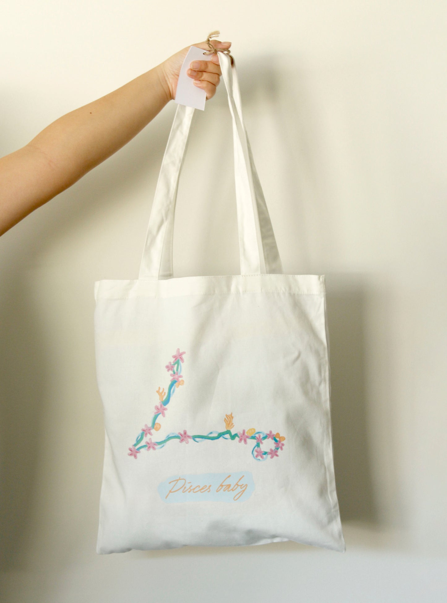 White tote bag with a Pisces astrology symbol on it, held by a hand against a light background