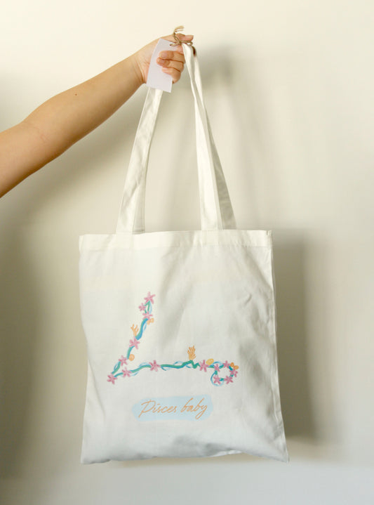 White tote bag with a Pisces astrology symbol on it, held by a hand against a light background