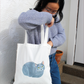 Person holding a tote bag with a russian blue cat design, standing outdoors.