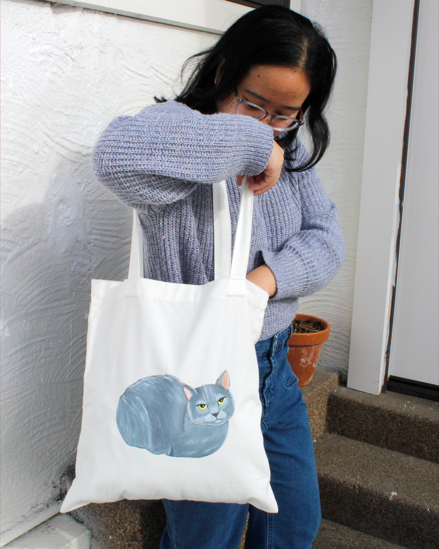 Person holding a tote bag with a russian blue cat design, standing outdoors.