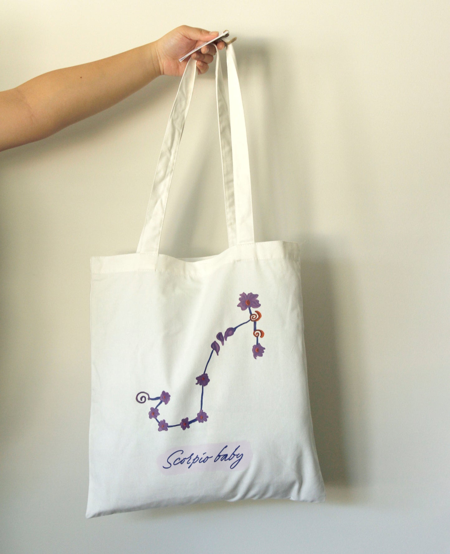 White tote bag with purple Scorpio zodiac design held by a hand against a light background