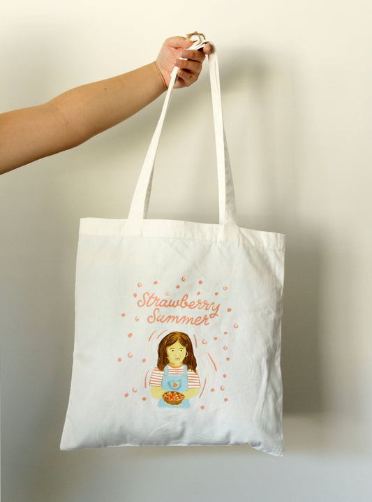 White tote bag with 'Strawberry Summer' design held by a hand against a plain background