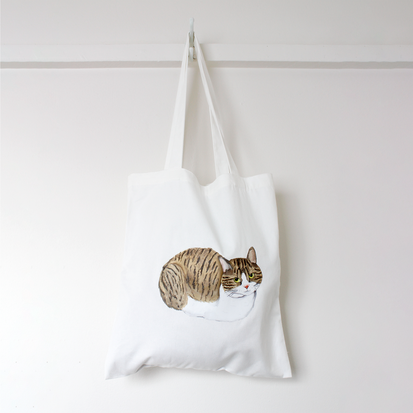 White tote bag with a tuxedo tabby cat loafing illustration hanging against a white background
