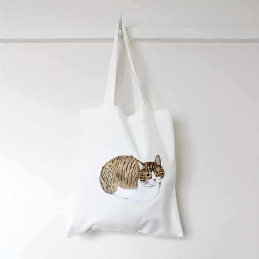 White tote bag with a tuxedo tabby cat loafing illustration hanging against a white background