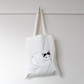 White tote bag with a cat illustration hanging on a white wall.