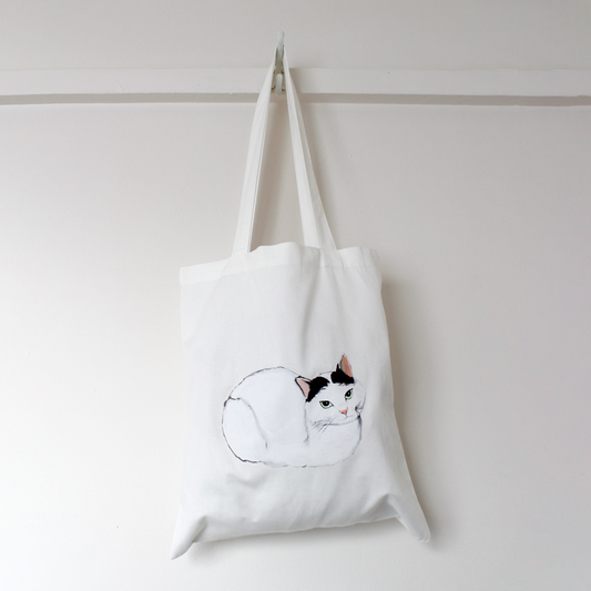 White tote bag with a cat illustration hanging on a white wall.