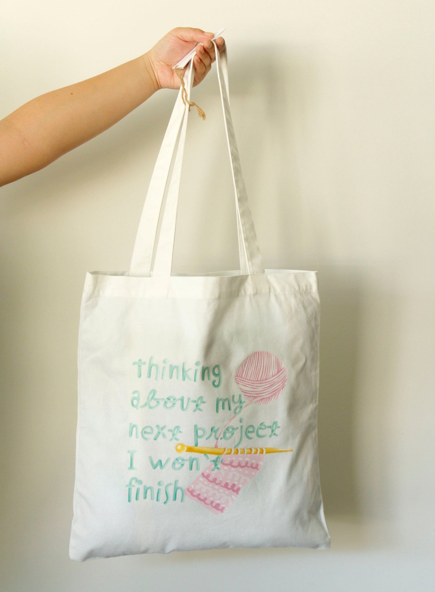 Tote bag with text and a yarn ball graphic held by a hand against a plain background